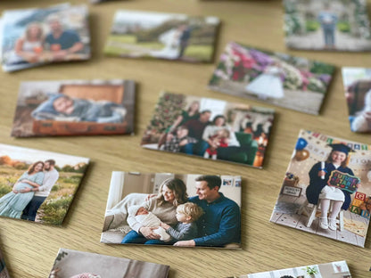 A close-up view of custom fridge magnets resting on a wooden surface, focusing on a magnet of a mother and father holding their newborn baby.