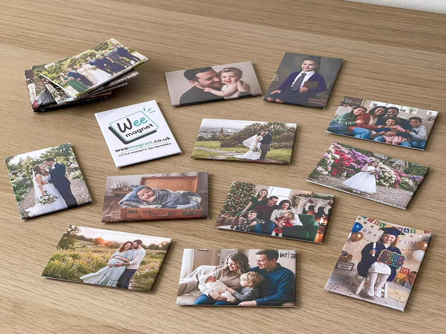 A wide variety of personalized Wee Magnet photo magnets scattered on a wooden table, featuring family portraits, newborn babies, weddings, and graduations.