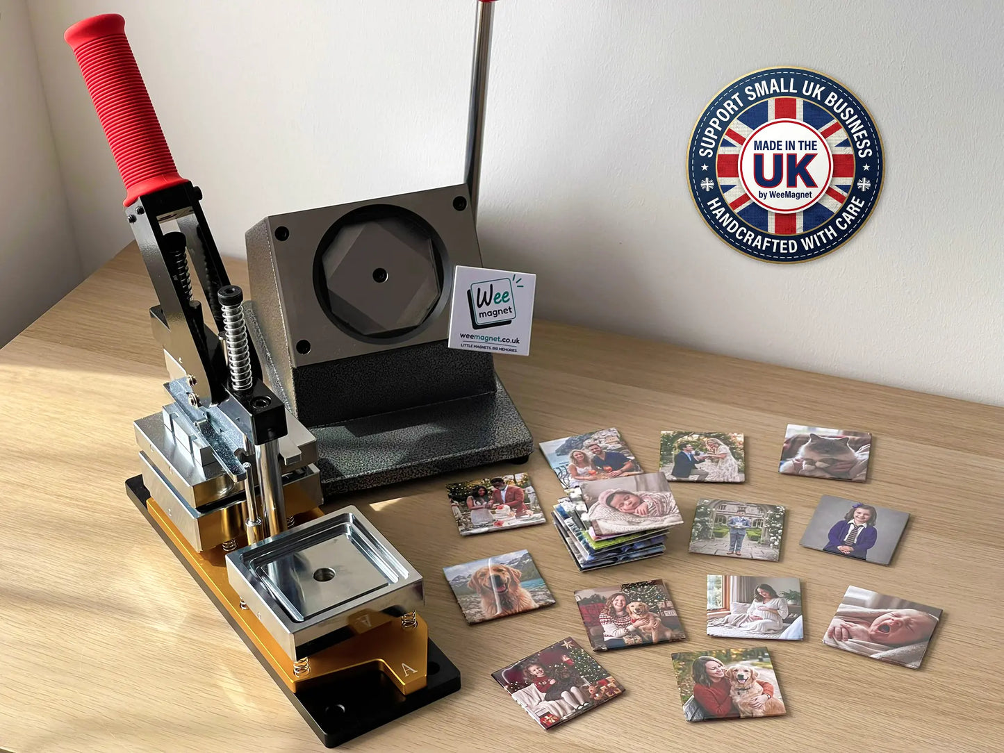 A heavy-duty magnet press machine sitting on a wooden desk next to a pile of freshly made square custom photo magnets, featuring a Made in the UK Support Small Business badge in the corner.