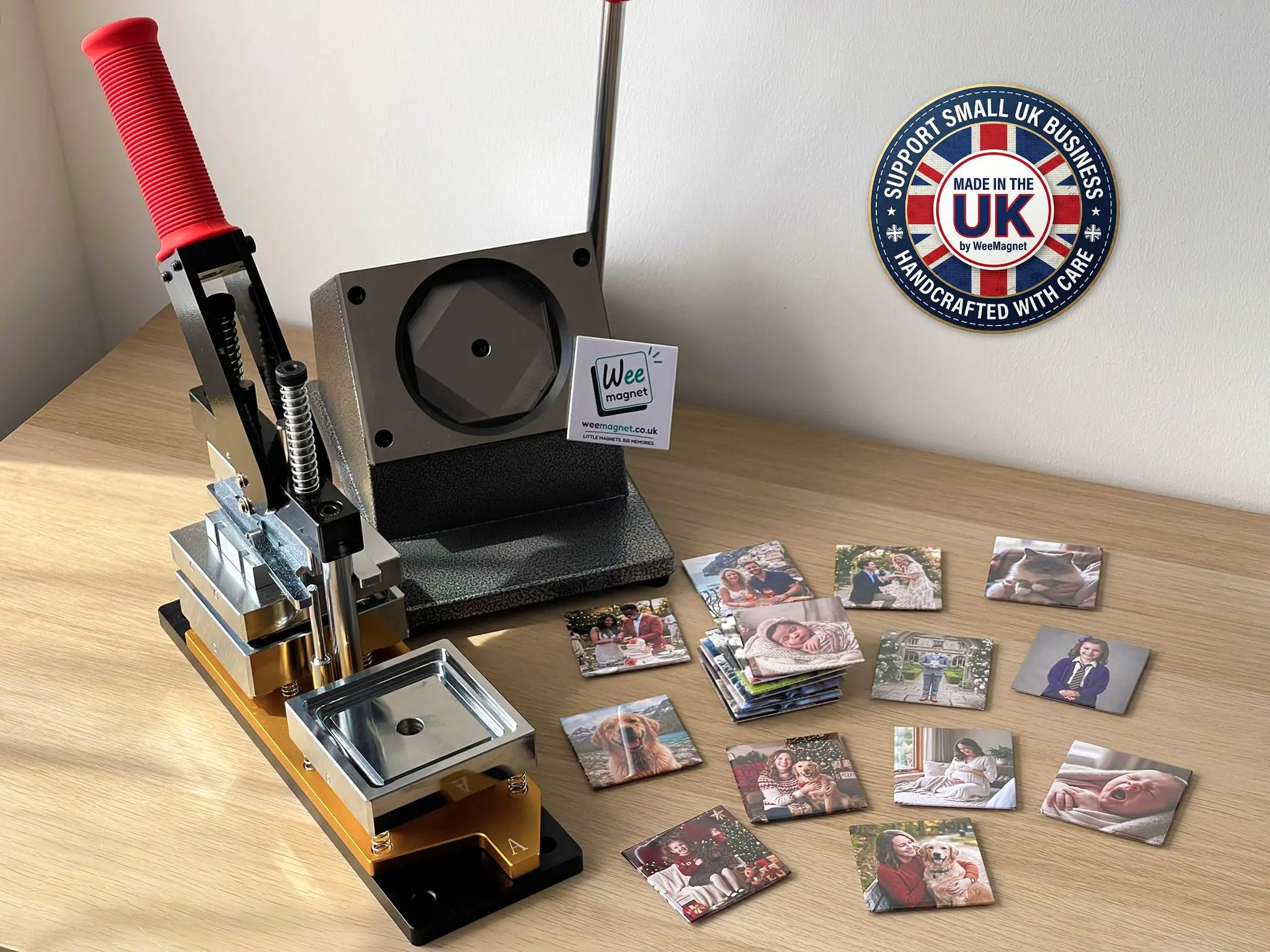 A heavy-duty magnet press machine sitting on a wooden desk next to a pile of freshly made square custom photo magnets, featuring a Made in the UK Support Small Business badge in the corner.
