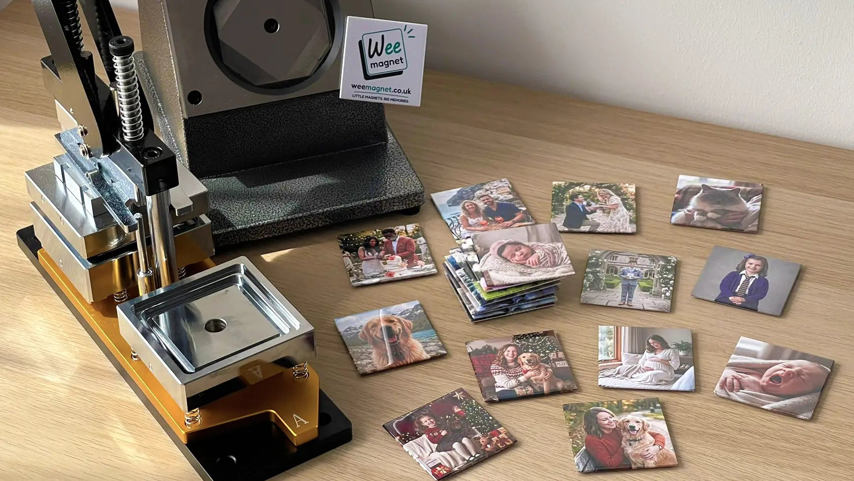 A handmade production setup on a light wooden desk, featuring a heavy-duty magnet press machine and a collection of freshly made square personalised photo magnets. The custom magnets, showcasing weddings, newborn babies, and family pets, are stacked and scattered next to a Wee Magnet logo magnet.